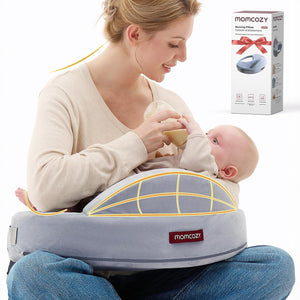 Mamá dándole mamadera a su bebé sobre un cojín de lactancia Momcozy gris con líneas que marcan la barrera de seguridad, junto a la caja del producto