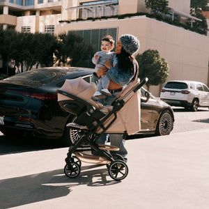 mujer con bebé en brazos junto a travel system beezy 2026 almond beige con silla de auto cloud g de cybex