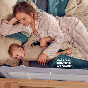 Mamá en pijama a rayas recostada junto a su bebé en una cama con ropa de cama verde azulada y almohadas en tonos neutros. Barrera gris con borde acolchado visible en el extremo inferior de la imagen, protegiendo el lateral de la cama- Mini Nuts - Expertos en sillas de auto y coches de paseo
