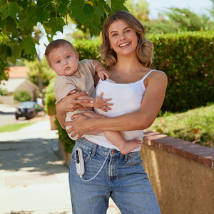 Mamá con su bebé en brazos extraendose leche con Extractor de leche materna Doble V2 Ultra ligero y potente de MOMCOZY mientras dan un paseo por la calle