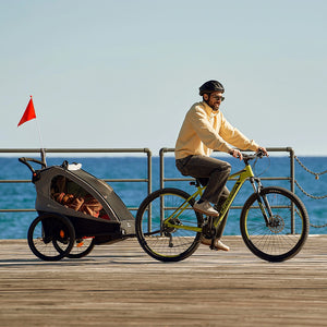 Hombre en bicicleta remolcando el Nava Walk 2 en 1 Kinderkraft por un paseo costero, en modo remolque de bicicleta