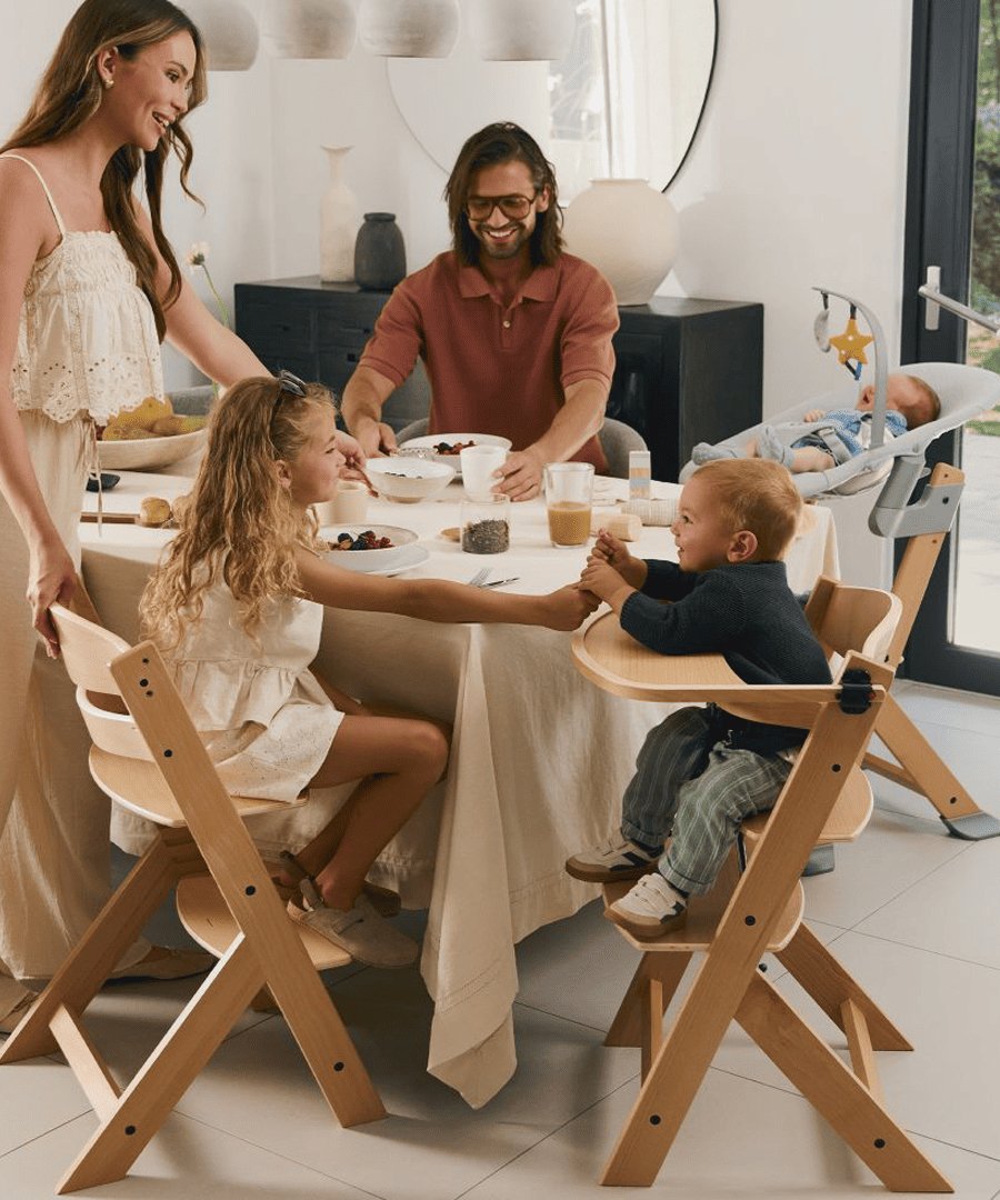 mamá parada frente a la mesa de comedor, papá sentado con un vaso en la mano, los niños en primer plano, una niña grande sentada en silla de comer enock madera pasandole un helado a un niño más pequeño que está sentado en silla de comer enock con bandeja y arnés