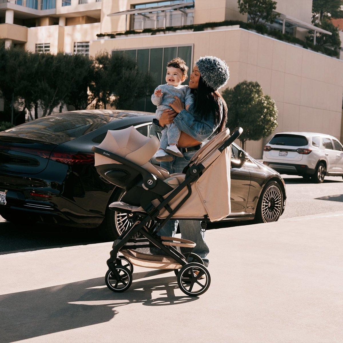 mujer con bebé en brazos junto a travel system beezy 2026 almond beige con silla de auto cloud g de cybex