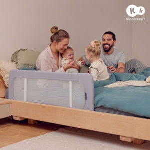 Mamá con bebé en brazos y papá sonriendo junto a niña pequeña sobre una cama baja de madera con ropa de cama verde azulada. Barrera gris con malla blanca instalada en el lateral de la cama, habitación cálida con suelo de madera

 - Mini Nuts - Expertos en sillas de auto y coches de paseo