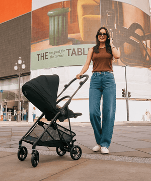 mujer en la calle de una ciudad posando junto a un coche de paseo Melio negro de cybex con vista lateral trasera 