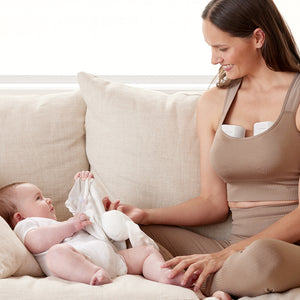 Extractor de leche materna S12 Pro Momcozy, madre usando el extractor bajo la ropa mientras sostiene a su bebé en el sofá