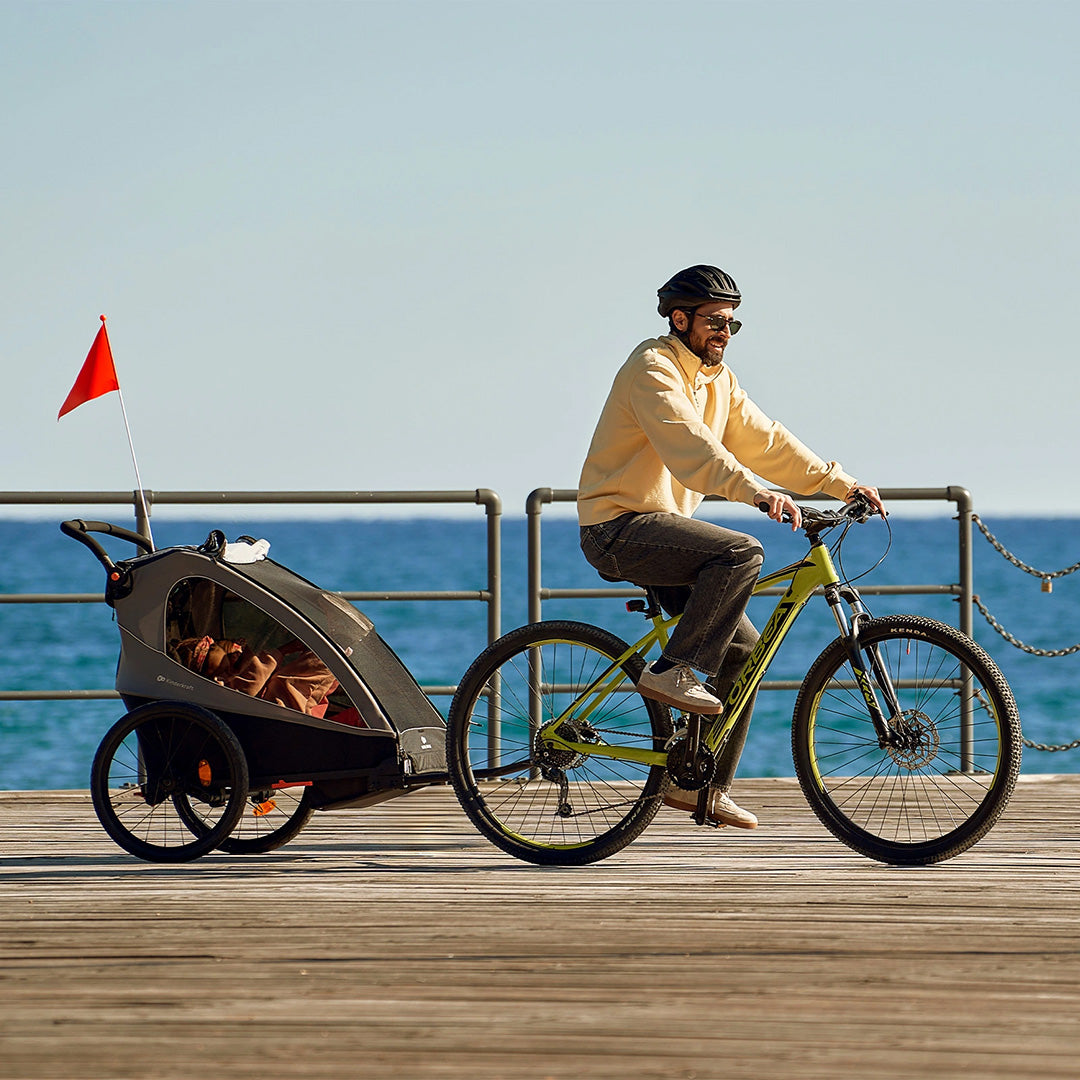 Hombre en bicicleta remolcando el Nava Walk 2 en 1 Kinderkraft por un paseo costero, en modo remolque de bicicleta