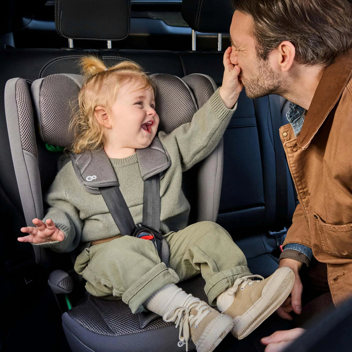 Niño pequeño sentado en la silla de auto Combinada Fix2Go i-Size Kinderkraft con el arnés de 5 puntas abrochado, tocando la cara de su papá mientras viajan en el auto